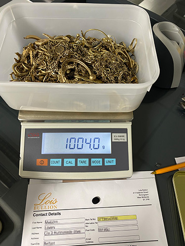 Tub of 9ct scrap gold weighing 1004 grams on digital scales next to a Lois Bullion seller form.