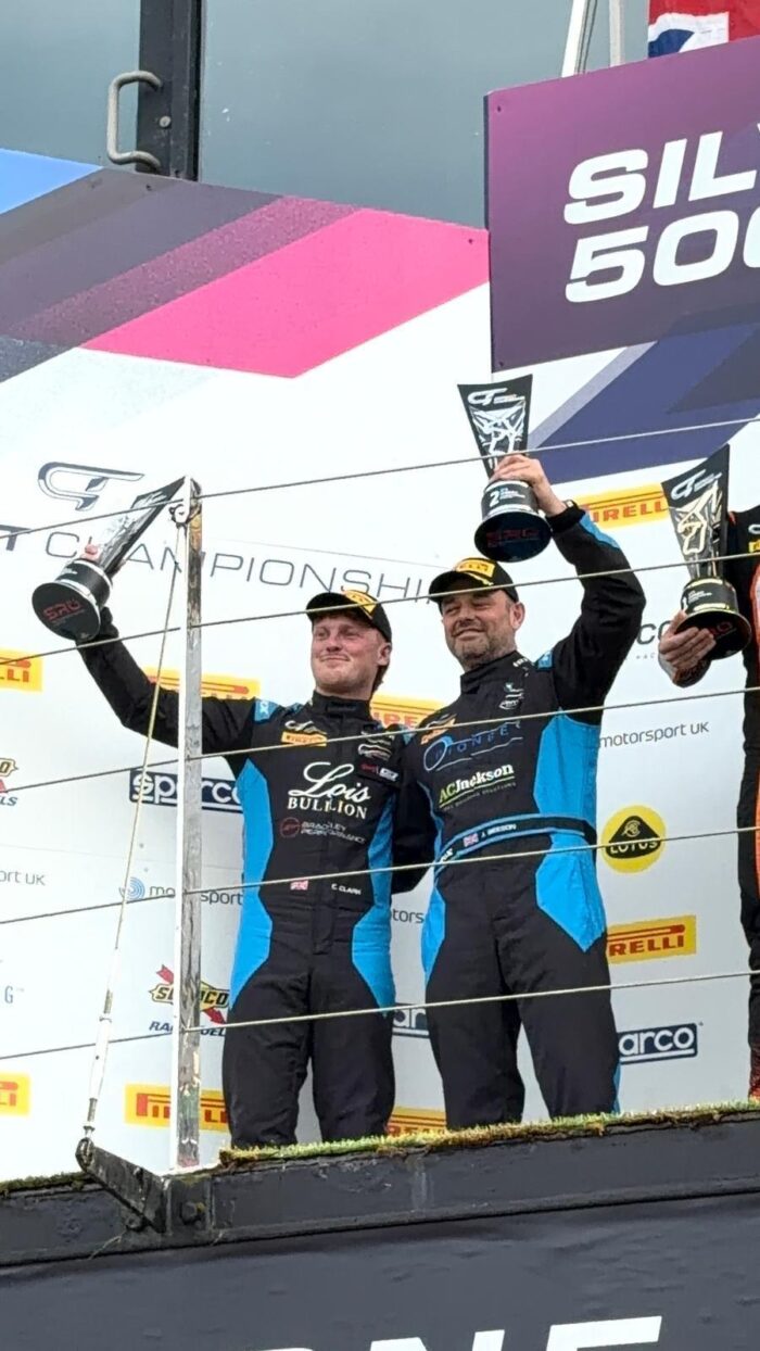 Charles Clark celebrating on the podium in a racing suit displaying the Lois Bullion logo