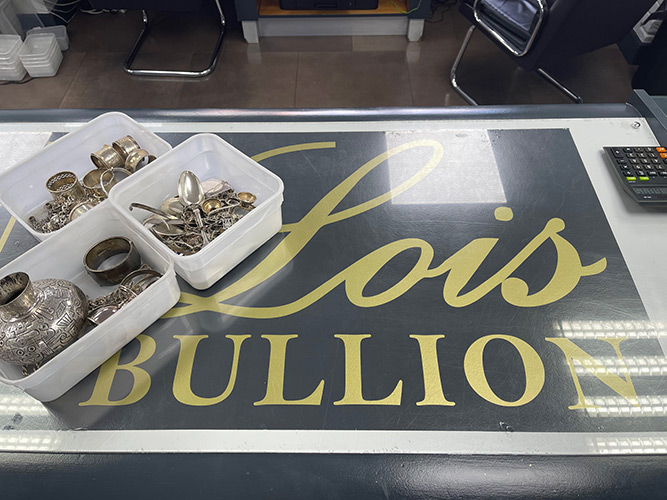 Lois Bullion counter with branding and tubs of scrap silver items including spoons, vases, napkin rings, candlestick holders, bracelets, and chains.