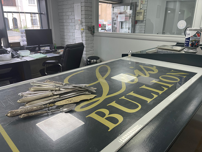 Lois Bullion counter with branding and scrap silver items including knives, fish knife, and butter knife.